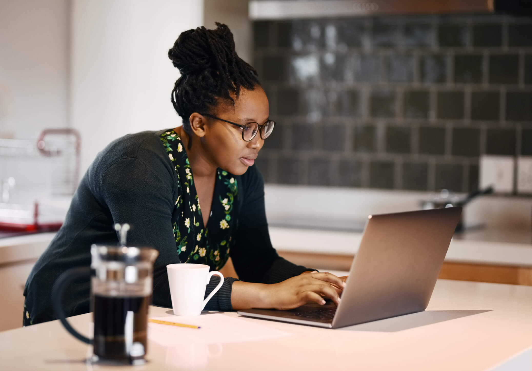 Mid-adult Black woman searching for information on her laptop. She is sitting at the kitchen island inside her home while drinking a cup of coffee.