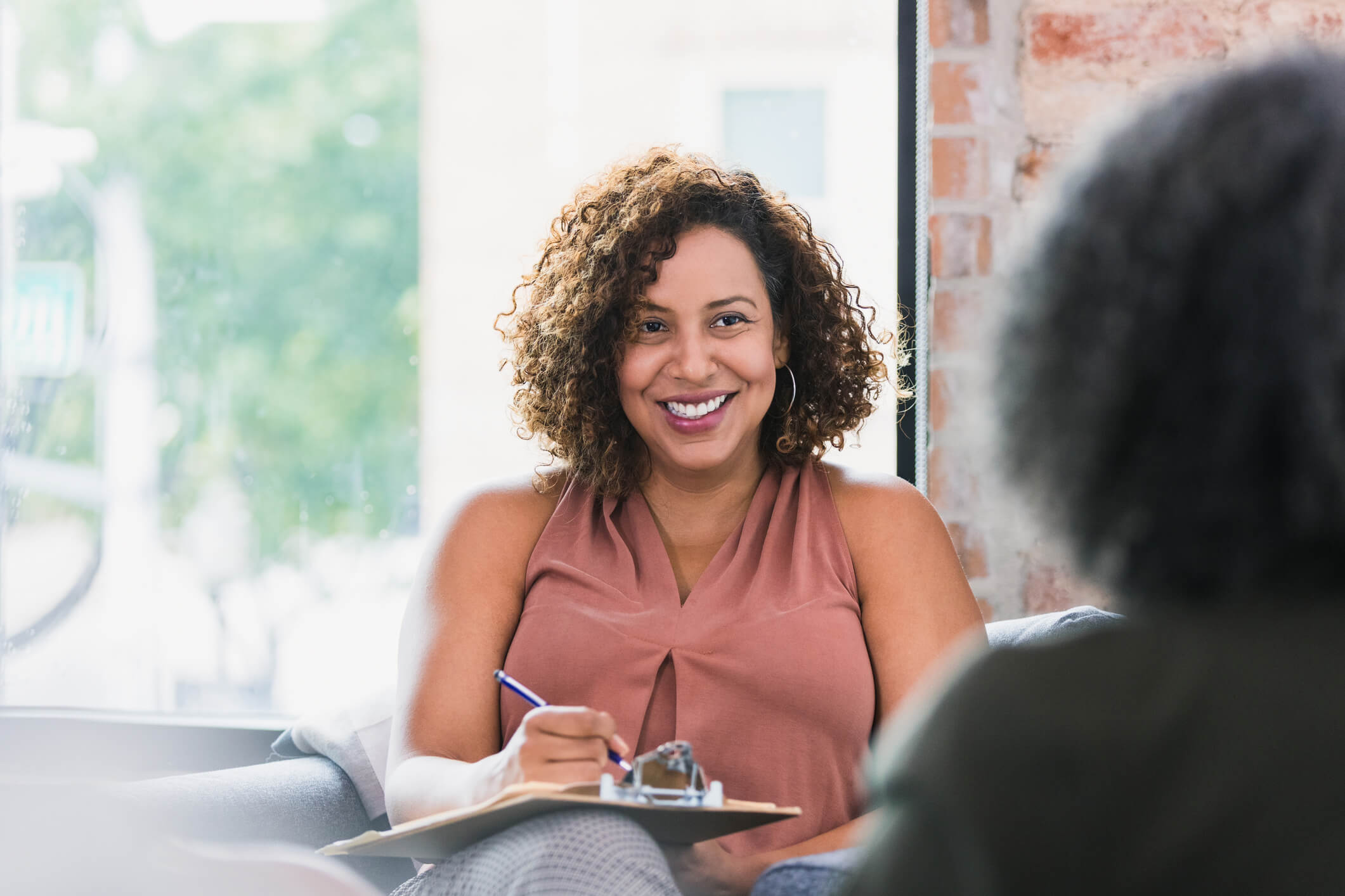 A smiling mid-adult female counselor sitting across from a female patient is taking notes.